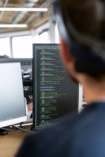 The image shows a close-up of a person working at a desk in an office environment, viewed from behind. The individual, with short dark hair, is focusing on a large computer monitor displaying lines of