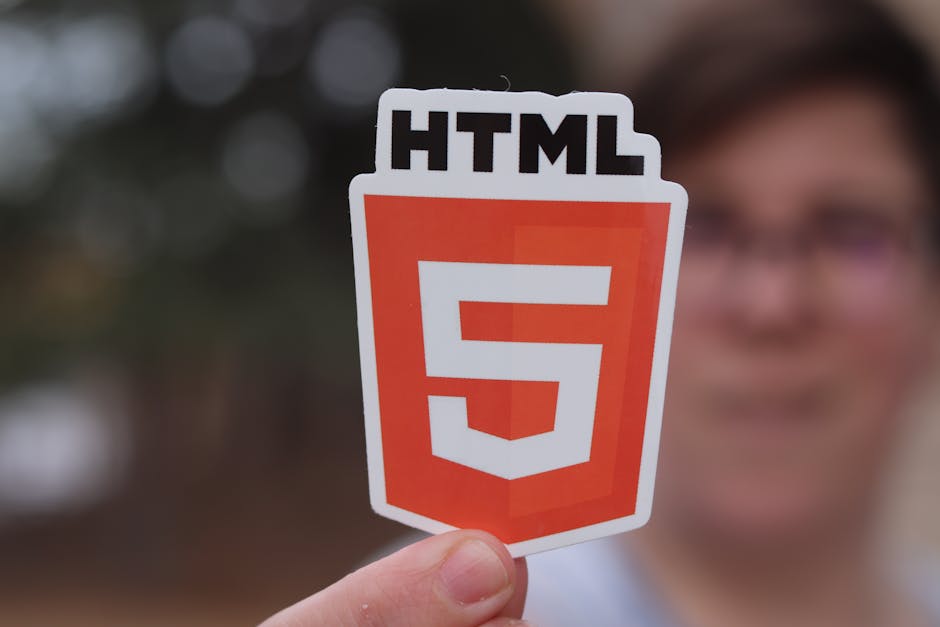 A person holding a small sticker featuring the HTML5 logo, which consists of an orange shield shape with a white number '5' in the center and the text 'HTML' at the top. The background is blurred, sho